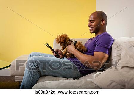 Stock Image - Mid adult male sitting on sofa with dog looking at digital tablet. Fotosearch