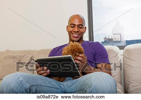 Stock Image - Mid adult male sitting on sofa with dog and digital tablet. Fotosearch