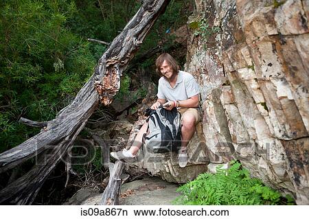 Picture - Portrait of young male hiker photographing sitting on rock. Fotosearch