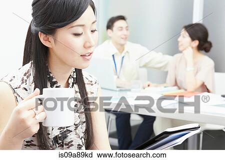 Stock Image - Woman holding mug, colleagues in background. Fotosearch