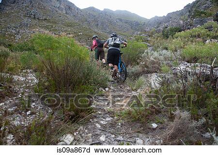 Stock Image - Young couple pushing mountain bikes up mountain. Fotosearch