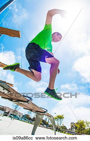 Stock Image - Young man leaping over park bench in city. Fotosearch