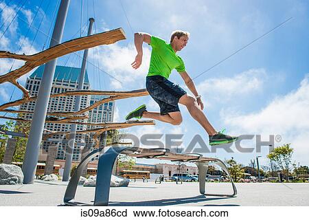 Stock Image - Young man leaping over park bench in city. Fotosearch