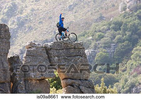 Stock Image - Young man on mountain bike standing on rock formation. Fotosearch