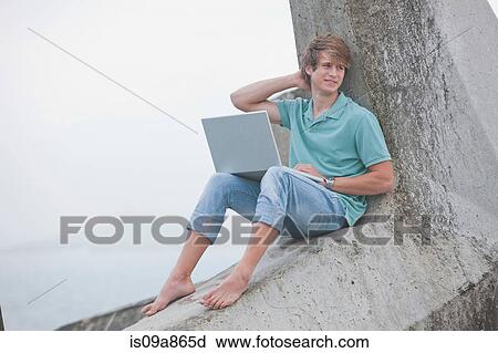 Stock Image - Young man using laptop on pier. Fotosearch