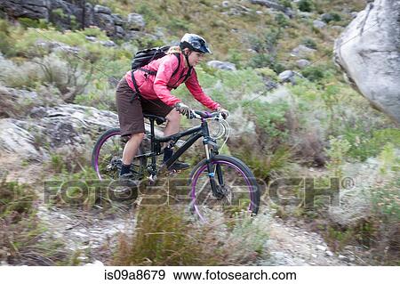 Stock Photo - Young woman mountain biking through scrubland. Fotosearch