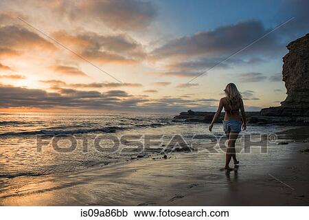 Stock Image - Young woman walking on beach at sunset, rear view. Fotosearch