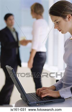 Stock Image - Woman using laptop as colleagues shake hands. Fotosearch