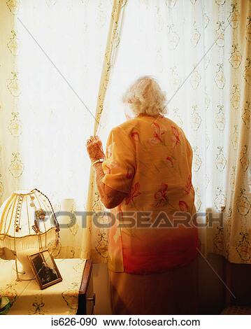 Stock Image - Elderly woman looking opening curtain. Fotosearch