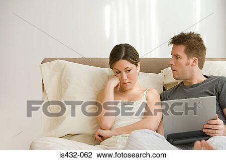 Stock Image - Wife annoyed at husband using laptop in bed. Fotosearch