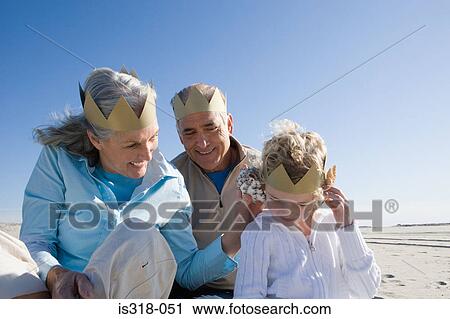 Stock Image - Grandparents and granddaughter wearing paper crowns on beach. Fotosearch