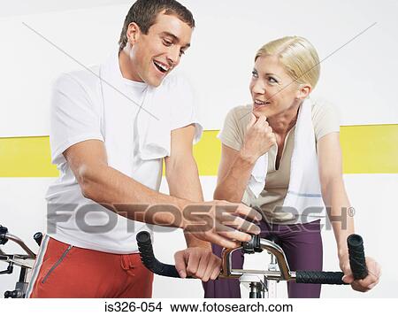 Picture - Woman sitting on exercise bike man checking control panel. Fotosearch