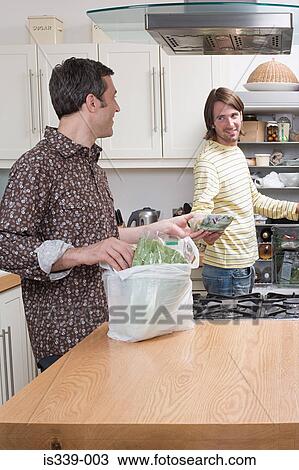 Stock Image - Gay couple putting food into fridge. Fotosearch