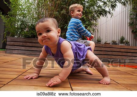 Stock Photo - Baby girl crawling on patio, brother in background. Fotosearch