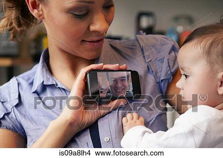Baby girl watching father on video call, mother holding smartphone View Large Photo Image Picture - Baby girl watching father on video call, mother holding smartphone. Fotosearch