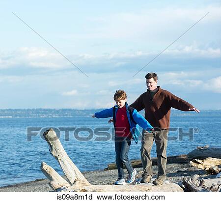 Stock Image - Father and son walking across tree trunk at coast. Fotosearch
