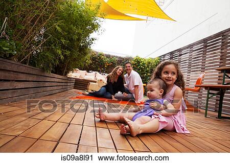 Stock Image - Girl with baby sister sitting on lap, parents in background. Fotosearch
