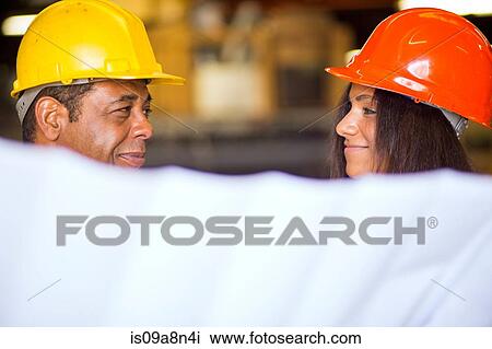 Stock Image - Man and woman in warehouse holding blueprint. Fotosearch