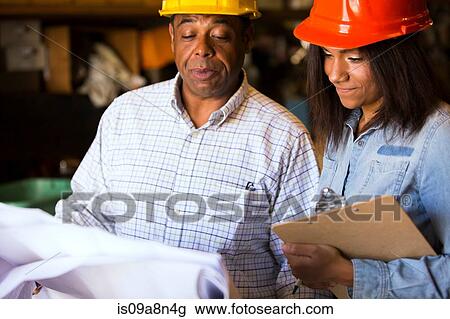 Stock Image - Man and woman in warehouse with blueprint and clipboard. Fotosearch