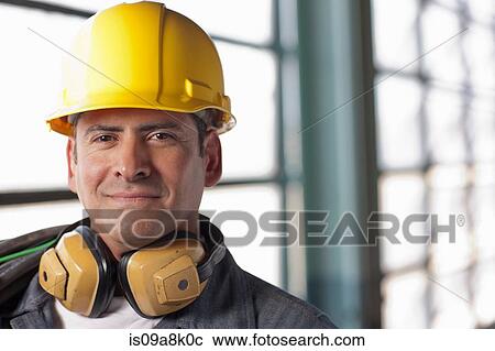 Stock Image - Mature construction worker wearing hard hat and ear defenders, smiling. Fotosearch