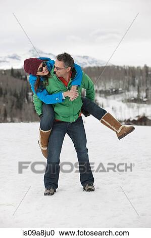 Stock Image - Mature man carrying young woman on back in snow. Fotosearch