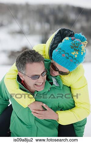 Stock Image - Mature man carrying young woman on back in snow, laughing. Fotosearch