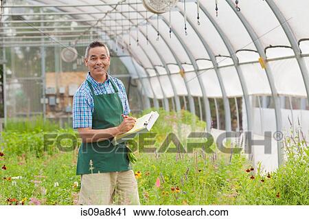 Stock Image - Mature man holding clipboard in garden centre, portrait. Fotosearch