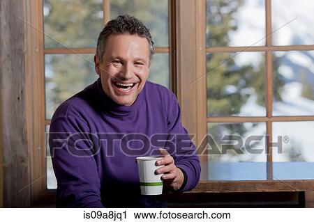 Stock Image - Mature man holding hot drink indoors, laughing. Fotosearch