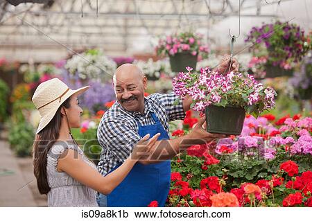 Stock Image - Mature man serving young woman in garden centre, smiling. Fotosearch