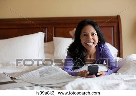 Stock Image - Mature woman lying on bed holding mobile phone, portrait. Fotosearch