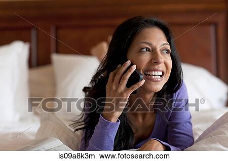 Picture - Mature woman lying on bed using mobile phone, smiling. Fotosearch