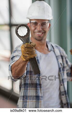Stock Image - Mid adult construction worker wearing hard hat and holding wrench, portrait. Fotosearch