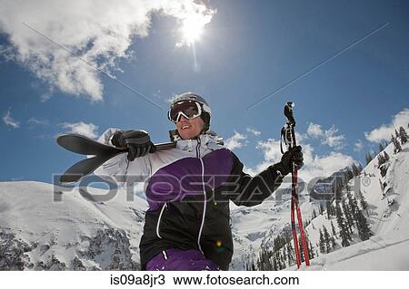 Stock Image - Mid adult man in skiwear carrying skis on shoulder, smiling. Fotosearch