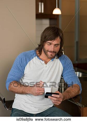 Stock Image - Mid adult man looking at mobile phone in kitchen. Fotosearch