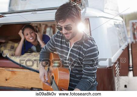 Stock Photo - Mid adult man playing guitar at back of camper van, smiling. Fotosearch