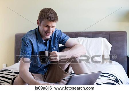 Stock Photograph - Mid adult man sitting on bed using laptop. Fotosearch