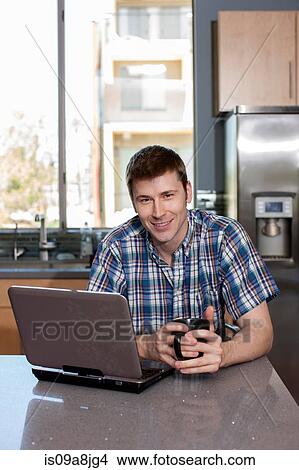 Picture - Mid adult man using laptop in kitchen, portrait. Fotosearch