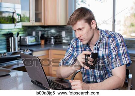 Stock Photography - Mid adult man using laptop in kitchen. Fotosearch