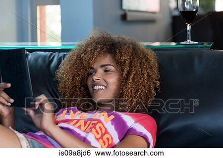 Stock Photograph - Mid adult woman relaxing on sofa with electronic book, smiling. Fotosearch