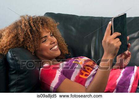 Stock Photography - Mid adult woman relaxing on sofa with electronic book, smiling. Fotosearch