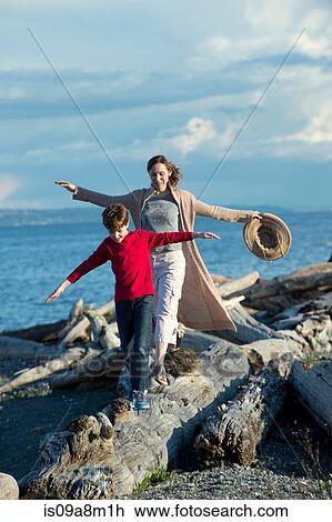 Stock Image - Mother and son walking along tree trunk at coast. Fotosearch