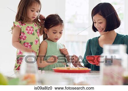 Stock Image - Mother and two daughters spooning topping onto tarts. Fotosearch