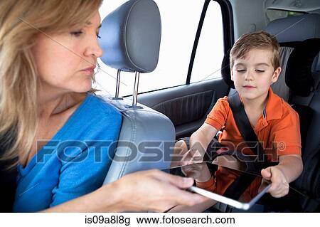 Stock Image - Mother passing digital tablet to son in back seat of car. Fotosearch