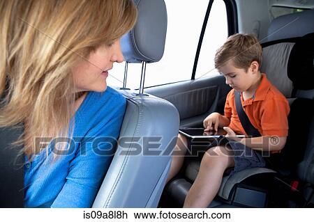 Stock Image - Mother watching son use digital tablet in back seat of car. Fotosearch