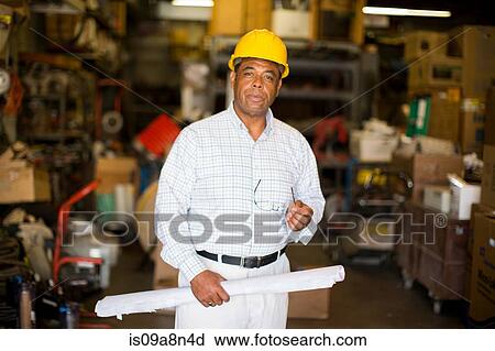 Stock Image - Portrait of man in warehouse holding blueprint. Fotosearch