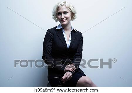 Stock Image - Studio portrait of blond businesswoman sitting on stool. Fotosearch
