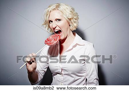 Stock Image - Studio portrait of mature woman biting into red lollipop. Fotosearch