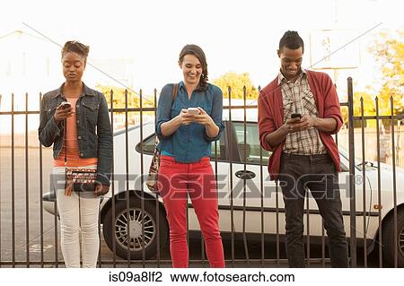 Stock Image - Three friends using cell phones by metal fence. Fotosearch