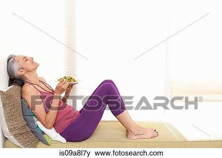 Stock Photo - Woman sitting on window seat eating lunch, head back. Fotosearch
