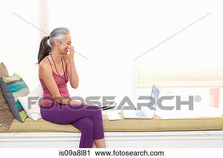 Stock Image - Woman sitting on window seat using laptop. Fotosearch